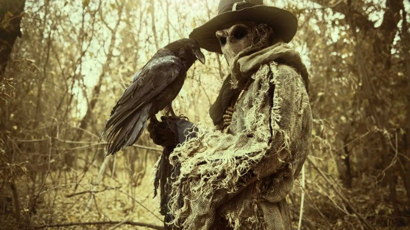 A terrible forest sorcerer with a canvas bag on his head and in a sackcloth robe stands in a dense forest with a black raven on his hand. Scarecrow. Halloween Tales. Horror, thriller.